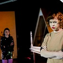 Na pierwszym planie kobieta z założonymi rękami patrzy przed siebie, na drugim planie druga kobieta na tle otwartych drzwi.