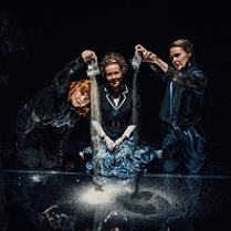 Trzy kobiety, dwie z nich wysypują piasek z woreczków na czarną powierzchnię. Czarne tło.