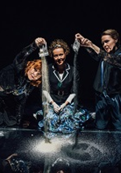 Trzy kobiety, dwie z nich wysypują piasek z woreczków na czarną powierzchnię. Czarne tło.