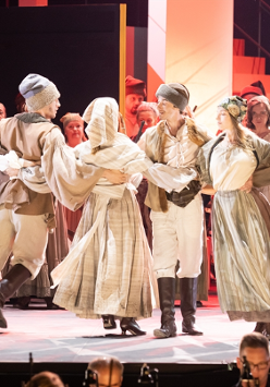 Grupa tancerzy w jasnych ludowych strojach tańczą trzymając się za ręce, w tle aktorzy, którzy patrzą na tańczących.