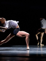 Tancerka w białej koszuli i czarnych szortach w tanecznej pozie - wygięta do tyłu, z jedną nogą zgięta, a drugą wyprostowaną. W tle druga osoba stoi z ugiętą nogą, czarne tło.