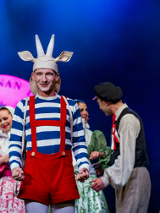 Aktor przebrany za Koziołka Matołka, w czerwonych spodniach na szelkach i biało-niebieskiej bluzce, za nim kilka osób w strojach ludowych.