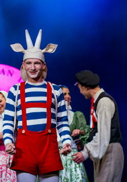 Aktor przebrany za Koziołka Matołka, w czerwonych spodniach na szelkach i biało-niebieskiej bluzce, za nim kilka osób w strojach ludowych.