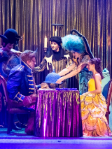 Przy stoliku siedzi kobieta w żółtej sukni, naprzeciw niej siedzi postawny mężczyzna w granatowym stroju, z rogami na głowie. Obok stołu stoją trzy postacie w strojach scenicznych, jedna z nich trzyma nad stolikiem półokrągłe naczynie.