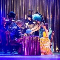 Przy stoliku siedzi kobieta w żółtej sukni, naprzeciw niej siedzi postawny mężczyzna w granatowym stroju, z rogami na głowie. Obok stołu stoją trzy postacie w strojach scenicznych, jedna z nich trzyma nad stolikiem półokrągłe naczynie.