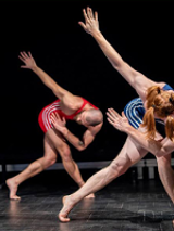 Dwoje aktorów w sportowych strojach na scenie, w takich samych pozach - pochyleni, ze skrzyżowanymi nogami i jedną ręką wyprostowaną ku górze.