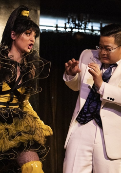 na zdjęciu mężczyzna ubrany w biały garnitur stoi unosząc ręce przed siebie przed krzyczącą na niego kobietą, ubraną w żółto-czarną sukienkę