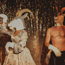 Dwie postacie na scenie: mężczyzna z nagim torsem i kobieta w sukni w srebrno-złotych kolorach. Aktorzy mają założone głowy kozłów. W tle mieniąca się tkanina.