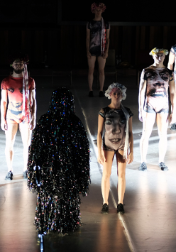 Kilka postaci stojących w pewnej odległości od siebie, w strojach z nadrukowanymi twarzami. Na pierwszym planie postać pokryta sznurami małych, świecących, kolorowych światełek.