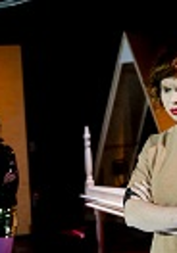 Na pierwszym planie kobieta z założonymi rękami patrzy przed siebie, na drugim planie druga kobieta na tle otwartych drzwi.