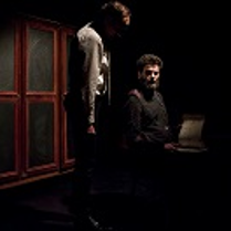 Ciemne zdjęcie: dwóch mężczyzn, jeden siedzi , drugi przy nim stoi. W tle drzwi szafy.