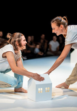 Dwie uśmiechnięte kobiety na scenie na przeciwko siebie, jedna stojąc pochyla się do przodu, druga kuca, obie dotykają białego, kartonowego domku. W tle publicznośc.