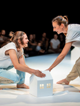 Dwie uśmiechnięte kobiety na scenie na przeciwko siebie, jedna stojąc pochyla się do przodu, druga kuca, obie dotykają białego, kartonowego domku. W tle publicznośc.