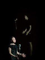 Kobieta w czarnej sukience stoi przed mikrofonem, za nią zdjęcie innej kobiety stojącej przed mikrofonem na czarnym tle.