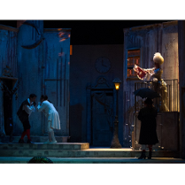 Na zdjeciu czworo aktorów w dziewiętnastowiecznych strojach na scenie. Po lewej stronie dwaj mężczyźni stoją pochyleni ku sobie, po prawej na balkonie widoczna kobieta w długiej sukni, przed nią na chodniku stoi mężczyzna w czarnym płaszczu i kapeluszu pod parasolem.