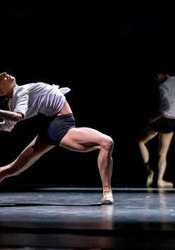 Tancerka w białej koszuli i czarnych szortach w tanecznej pozie - wygięta do tyłu, z jedną nogą zgięta, a drugą wyprostowaną. W tle druga osoba stoi z ugiętą nogą, czarne tło.