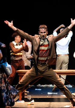 Mężczyzna z uniesionymi i wyprostowanymi rękoma, stojący w rozkroku, za nim drewniana ławka bez oparcia i kilku innych mężczyzn w różnych pozach.