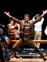 Mężczyzna z uniesionymi i wyprostowanymi rękoma, stojący w rozkroku, za nim drewniana ławka bez oparcia i kilku innych mężczyzn w różnych pozach.