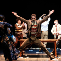 Mężczyzna z uniesionymi i wyprostowanymi rękoma, stojący w rozkroku, za nim drewniana ławka bez oparcia i kilku innych mężczyzn w różnych pozach.