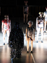 Kilka postaci stojących w pewnej odległości od siebie, w strojach z nadrukowanymi twarzami. Na pierwszym planie postać pokryta sznurami małych, świecących, kolorowych światełek.