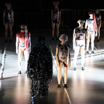 Kilka postaci stojących w pewnej odległości od siebie, w strojach z nadrukowanymi twarzami. Na pierwszym planie postać pokryta sznurami małych, świecących, kolorowych światełek.