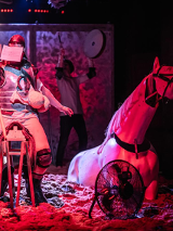 Mężczyzna z zakrytą twarzą siedzi na drewnianym siedzisku przypominającym siodło, trzyma w ręce długi kij. Po prawej stronie figura konia do połowy zakopana w piasku.