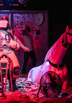 Mężczyzna z zakrytą twarzą siedzi na drewnianym siedzisku przypominającym siodło, trzyma w ręce długi kij. Po prawej stronie figura konia do połowy zakopana w piasku.