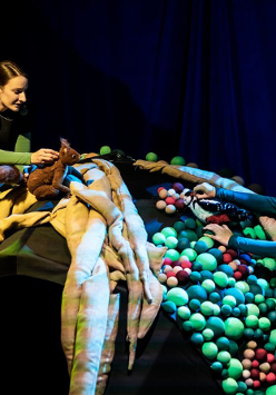 Na zdjęciu dwie kobiety, jedna trzyma pluszową wiewiórkę, druga - dzięcioła; obie aktorki stoją po dwóch stronach półkolistej konstrukcji złożonej z pasów materiału i kolorowych kulek.