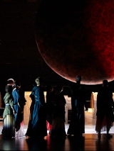 Ciemne zdjęcie sceny, nad którą unosi się duże czerwono-czarne koło przypominające księżyc. Na scenie aktorzy odwróceni tyłem do widzów i zwróceni w kierunku czerwonego koła. W tle elementy scenografii.