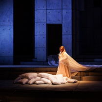 Oświetlona kobieta w długiej, białej szacie na scenie stoi przy położonych na ziemi kilkunastu białych poduszkach. Reszta sceny w kolorach czarno-niebieskich.