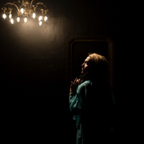 Ciemne zdjęcie, na którym widać kobietę odwróconą bokiem, z oświetloną twarzą. U góry zdjęcia świecąca się lampa w formie ozdobnego żyrandola.