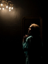 Ciemne zdjęcie, na którym widać kobietę odwróconą bokiem, z oświetloną twarzą. U góry zdjęcia świecąca się lampa w formie ozdobnego żyrandola.