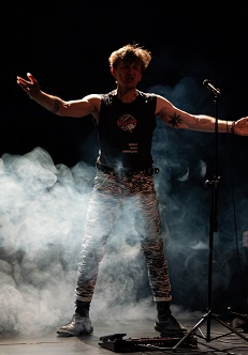 Mężczyzna stojący przy mikrofonie z rozpostartymi rękoma. Z boku stoi na stojaku gitara. Zdjęcie w półmroku, mężczyznę częściowo otacza biały dym.