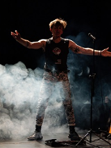 Mężczyzna stojący przy mikrofonie z rozpostartymi rękoma. Z boku stoi na stojaku gitara. Zdjęcie w półmroku, mężczyznę częściowo otacza biały dym.