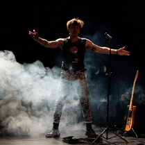 Mężczyzna stojący przy mikrofonie z rozpostartymi rękoma. Z boku stoi na stojaku gitara. Zdjęcie w półmroku, mężczyznę częściowo otacza biały dym.