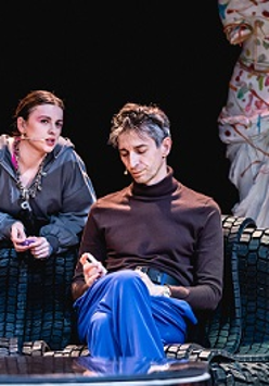 Mężczyzna siedzi na kanapie, za kanapą kobieta, która jest o nią oparta i skierowana w stronę mężczyzny. W tle fragment manekinu, na którym wisi kolorowa suknia.