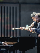 Na scenie trzech mężczyzn: jeden gra na fortepianie, dwaj pozostali stoją przy fortepianie zwróceni w kierunku pianisty. Jeden ze stojacych mężczyzn trzyma w dłoni pałeczke do gry na bębnach.