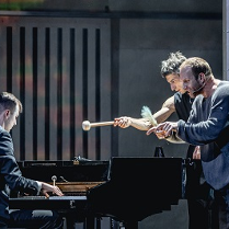 Na scenie trzech mężczyzn: jeden gra na fortepianie, dwaj pozostali stoją przy fortepianie zwróceni w kierunku pianisty. Jeden ze stojacych mężczyzn trzyma w dłoni pałeczke do gry na bębnach.