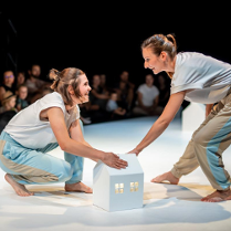 Dwie uśmiechnięte kobiety na scenie na przeciwko siebie, jedna stojąc pochyla się do przodu, druga kuca, obie dotykają białego, kartonowego domku. W tle publicznośc.