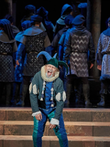 Błazen w zielono-niebieskim stroju i błazeńskiej czapce na scenie, za nim odwróceni tyłem do niego aktorzy, wszyscy w podobnych strojach i nakryciach głowy.