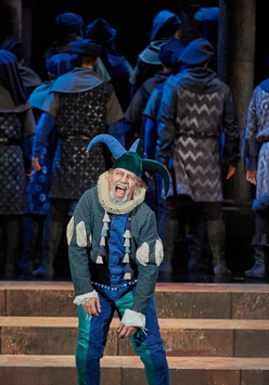 Błazen w zielono-niebieskim stroju i błazeńskiej czapce na scenie, za nim odwróceni tyłem do niego aktorzy, wszyscy w podobnych strojach i nakryciach głowy.