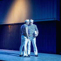 Na scenie dwóch mężczyzn stojących blisko siebie, w strojach przypominających marynarskie. Zdjęcie w niebiesko-błękitnych kolorach.