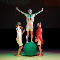 Trzech tancerzy, jeden z nich stoi z wyprostowanymi i uniesionymi do góry rękoma na dużej, zielonej piłce, pozostali stoją przy piłce bokiem do widza.