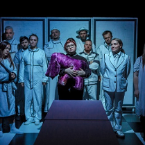 Na środku kobieta trzymająca fioletową przytulankę, za nią stoi dziesięć osób w białych ubraniach. Zdjęcie w niebiesko-białych kolorach.