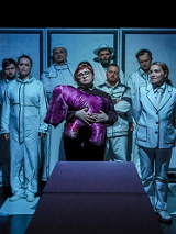 Na środku kobieta trzymająca fioletową przytulankę, za nią stoi dziesięć osób w białych ubraniach. Zdjęcie w niebiesko-białych kolorach.