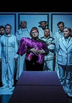 Na środku kobieta trzymająca fioletową przytulankę, za nią stoi dziesięć osób w białych ubraniach. Zdjęcie w niebiesko-białych kolorach.