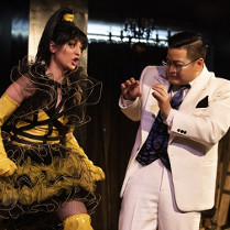 na zdjęciu mężczyzna ubrany w biały garnitur stoi unosząc ręce przed siebie przed krzyczącą na niego kobietą, ubraną w żółto-czarną sukienkę