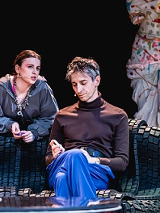 Mężczyzna siedzi na kanapie, za kanapą kobieta, która jest o nią oparta i skierowana w stronę mężczyzny. W tle fragment manekinu, na którym wisi kolorowa suknia.