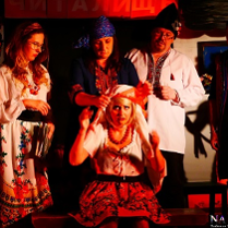 Aktorzy w strojach ludowych: jedna kobieta siedzi, druga nakłada jej białą chustę na głowę. Pozostałe osoby obserwują to.