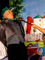 Trójka aktorów w strojach scenicznych (między innymi kucharza i smoka), w tle rysunek zamku.